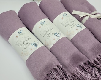 May include: Four rolled lavender shawls with fringed edges, each wrapped with a label reading "Thank You Claire's Bridal Shower 01.01.2024". One is tied with a white ribbon. These shawls are likely for a bridal shower.