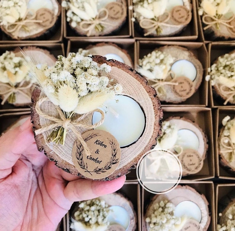 K&ouml;nnte beinhalten: Ein runder Kerzenhalter aus einer Holzscheibe mit einem wei&szlig;en Teelicht, einem kleinen Strau&szlig; aus getrockneten wei&szlig;en Blumen und Gr&auml;sern sowie einem runden Kraftpapieranh&auml;nger mit dem Text K&uuml;bra & Cankat, verpackt in einer braunen Pappschachtel.