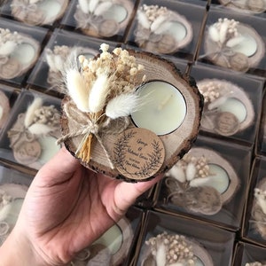 May include: A round wood slice candle holder featuring a white tea light, a small bouquet of dried cream-colored flowers and bunny tails, and a circular brown paper tag printed with the text Gamze & Mehmet Emin 19.08.2021 Nişan Hatırası.