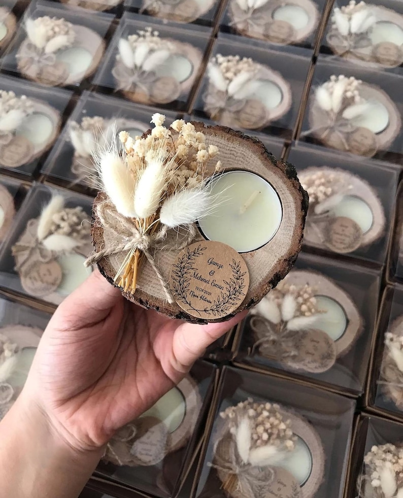 May include: A round wood slice candle holder featuring a white tea light, a small bouquet of dried cream-colored flowers and bunny tails, and a circular brown paper tag printed with the text Gamze & Mehmet Emin 19.08.2021 Nişan Hatırası.