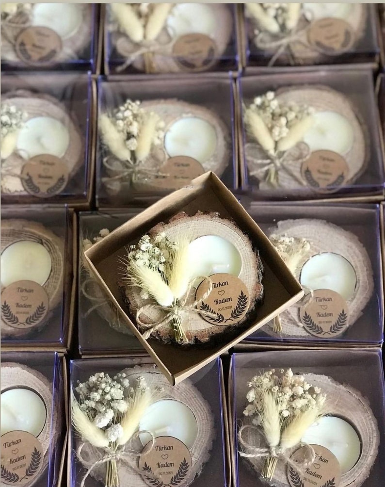 May include: A round wooden slice candle holder with a white tea light candle, decorated with a small bouquet of dried white flowers and grasses. A circular brown paper tag is attached, printed with the text T&uuml;rkan, a heart, Kadem, and 20.11.2021.