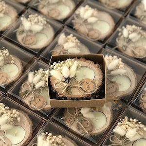 K&ouml;nnte beinhalten: Ein runder Kerzenhalter aus einer Holzscheibe mit einem wei&szlig;en Teelicht, verziert mit einem kleinen Strau&szlig; aus getrockneten wei&szlig;en Blumen und Gr&auml;sern, mit Bindfaden gebunden und einem runden Papieranh&auml;nger mit dem Aufdruck Melis & Şakir.