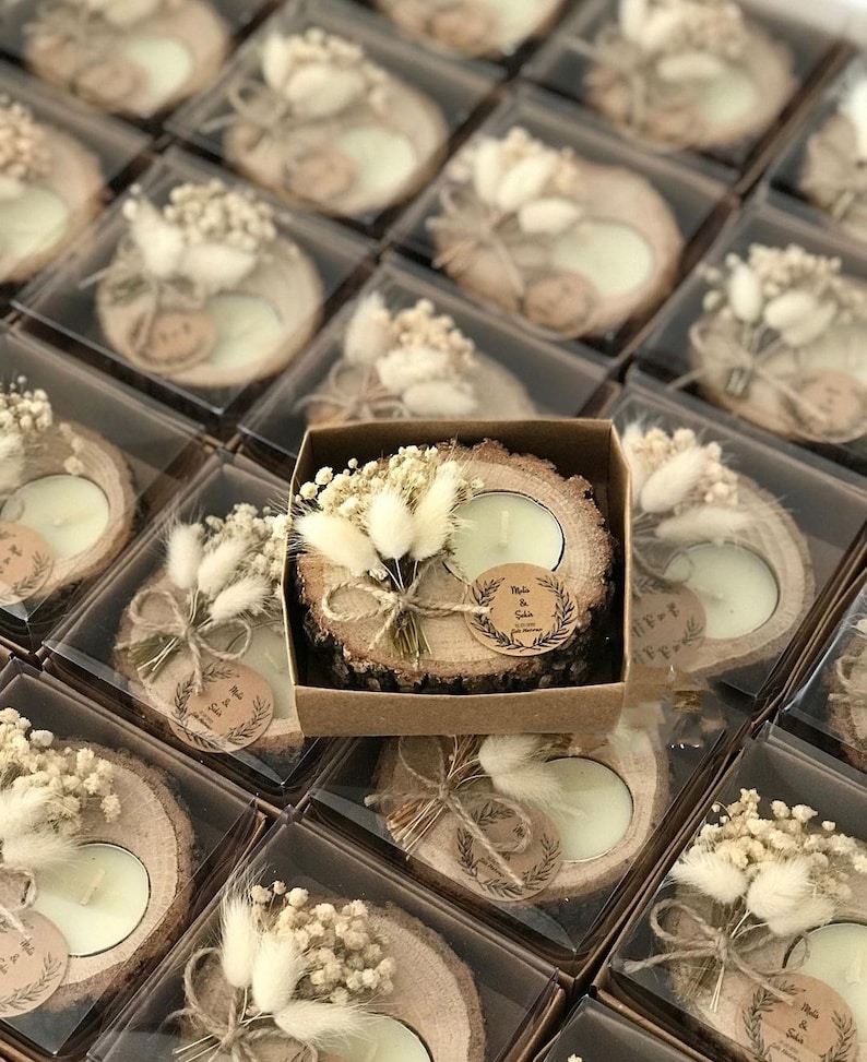 K&ouml;nnte beinhalten: Ein runder Kerzenhalter aus einer Holzscheibe mit einem wei&szlig;en Teelicht, verziert mit einem kleinen Strau&szlig; aus getrockneten wei&szlig;en Blumen und Gr&auml;sern, mit Bindfaden gebunden und einem runden Papieranh&auml;nger mit dem Aufdruck Melis & Şakir.