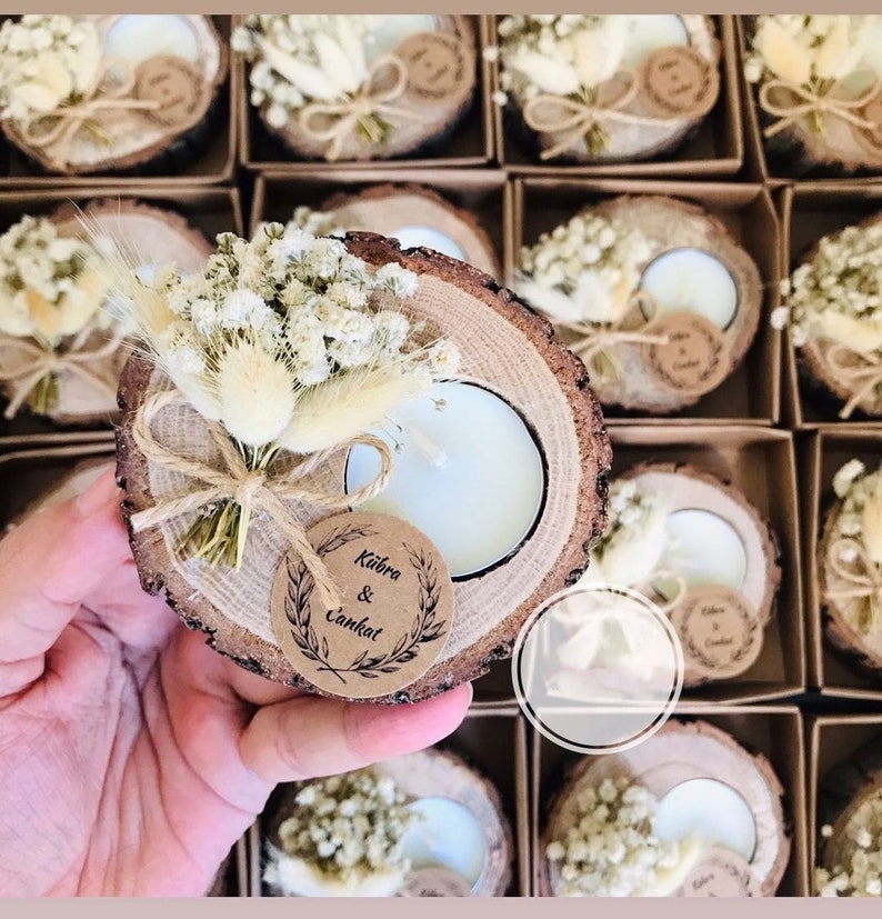 K&ouml;nnte beinhalten: Ein runder Kerzenhalter aus einer Holzscheibe mit einem wei&szlig;en Teelicht, einem kleinen Strau&szlig; aus getrockneten wei&szlig;en Blumen und Gr&auml;sern sowie einem runden braunen Papieranh&auml;nger, der mit einer Schnur befestigt ist. Der Anh&auml;nger tr&auml;gt den Text K&uuml;bra & Cankat.