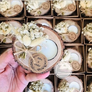 May include: A round wood slice candle holder featuring a white tea light candle, a small bouquet of dried white flowers and tan grasses, and a circular kraft paper tag printed with the names K&uuml;bra & Cankat, all tied together with twine.