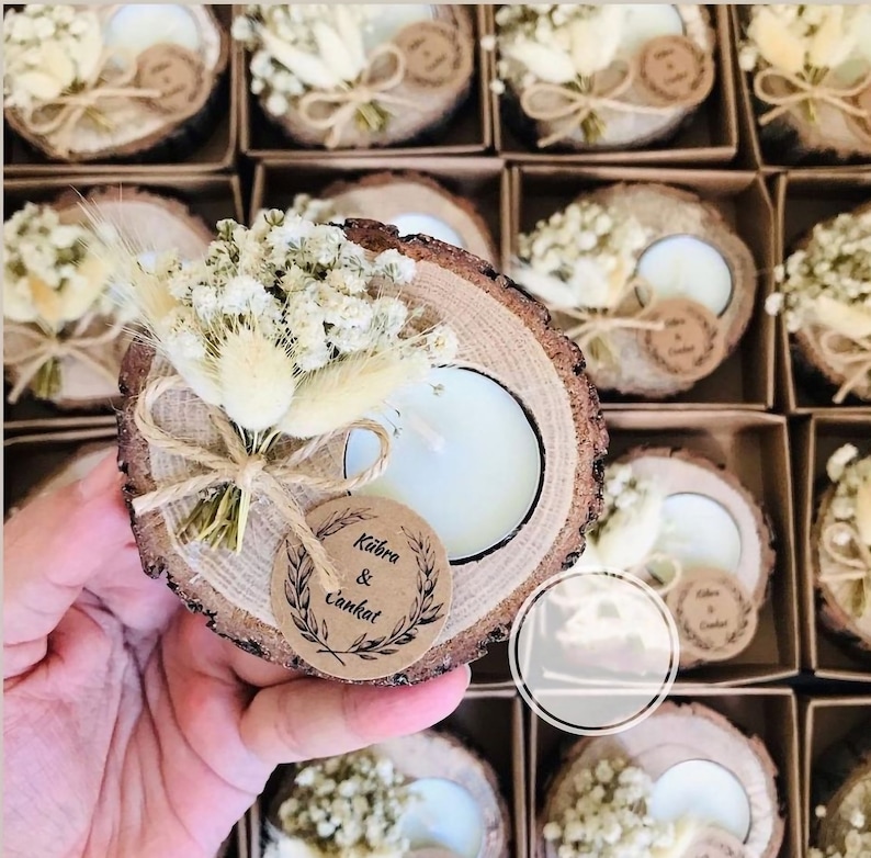 May include: A round wood slice candle holder featuring a white tea light candle, a small bouquet of dried white flowers and tan grasses, and a circular kraft paper tag printed with the names K&uuml;bra & Cankat, all tied together with twine.