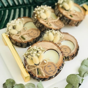 May include: Four round wood slice candle holders with natural bark edges, each featuring a white tea light candle, a small bouquet of dried white flowers and beige bunny tails, and a circular kraft paper tag printed with the text K&uuml;bra & Cankat.