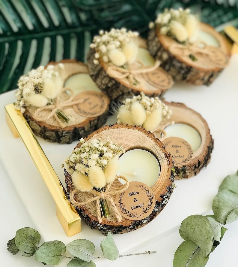 May include: Four round wood slice candle holders with natural bark edges, each featuring a white tea light candle, a small bouquet of dried white flowers and beige bunny tails, and a circular kraft paper tag printed with the text K&uuml;bra & Cankat.