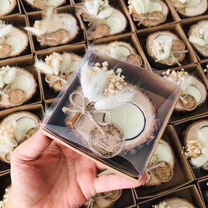 May include: A tea light candle set in a natural wood slice base, decorated with dried white bunny tail grass and baby's breath. The candle is encased in a clear plastic box with a round paper tag printed with the text Ebru & T&uuml;rker 10.07.2021.
