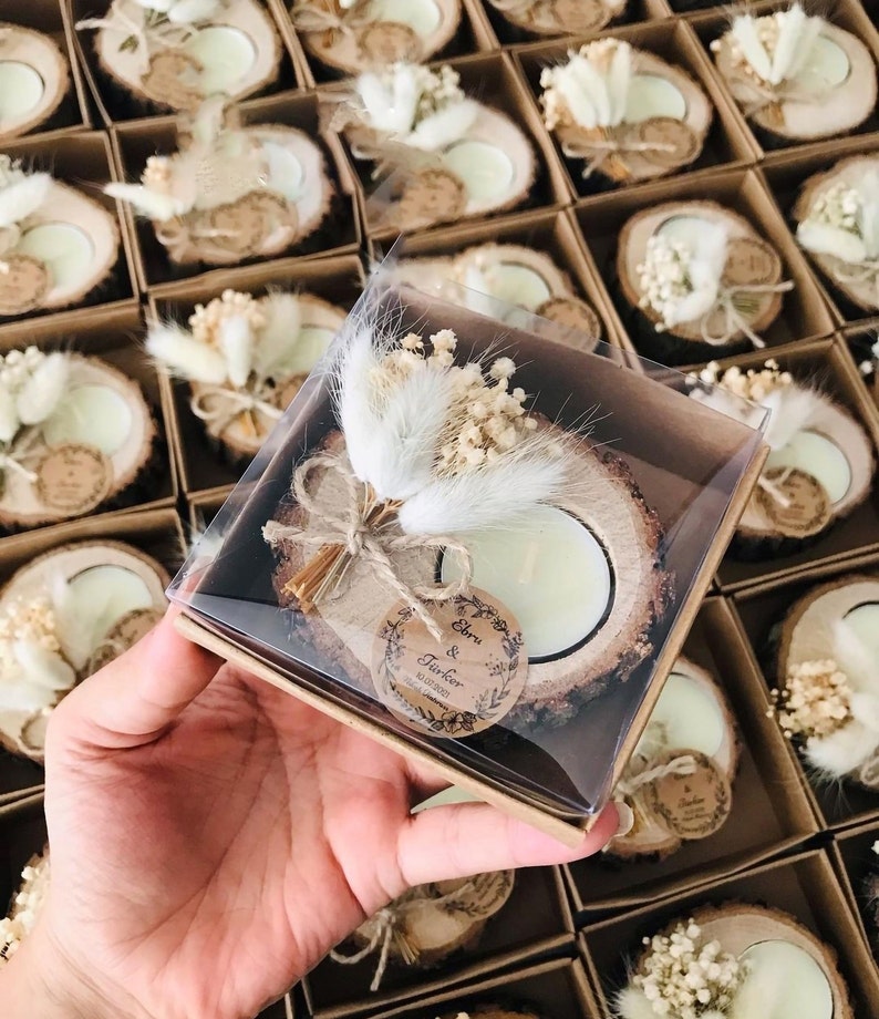 May include: A tea light candle set in a natural wood slice base, decorated with dried white bunny tail grass and baby's breath. The candle is encased in a clear plastic box with a round paper tag printed with the text Ebru & T&uuml;rker 10.07.2021.