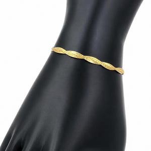 Peut inclure: Un bracelet de couleur dorée présentant un design torsadé et texturé. La bande métallique possède un motif entrelacé répétitif qui crée un effet de spirale subtil sur toute sa longueur, offrant une surface réfléchissante qui capte la lumière.