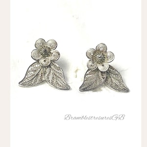 May include: A pair of silver-toned filigree earrings featuring a five-petaled flower design with two leaves extending from the base. The intricate metalwork creates a delicate, lace-like pattern throughout the floral and leaf shapes. Text reads BramblestresuresGB.