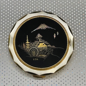 May include: A vintage compact mirror with a black enamel background and gold accents. The design features a Japanese mountain scene with a pagoda, a house, and a water wheel. The compact is marked with the letters "K24".