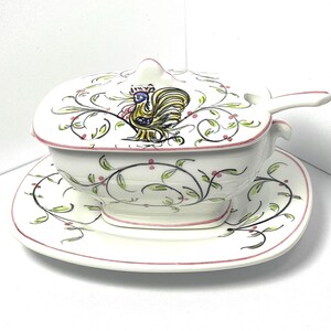May include: A white ceramic gravy boat with a matching lid, saucer, and ladle. The set features a hand-painted design of a yellow and blue cockerel on the lid, surrounded by delicate green vines and small pink berries, with pink trim edges.