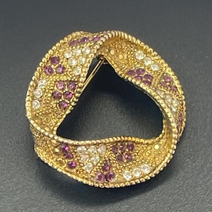 Pode incluir: Um broche circular em tom dourado com um design torcido semelhante a uma fita. A superfície apresenta uma borda texturizada com contas e é incrustada com grupos de pequenas pedras redondas, brancas transparentes e vermelho-rosa escuro.