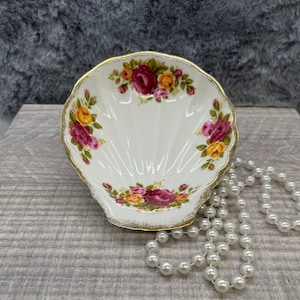 May include: A white porcelain shell-shaped dish with a gold rim and floral pattern. The dish is decorated with red, yellow, and pink roses. A string of white pearls is lying next to the dish.