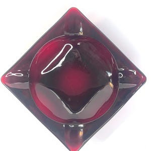 May include: A vintage ruby red glass ashtray. The ashtray is diamond-shaped with a circular center and three small rests for cigarettes. The glass has a glossy finish and reflects light.