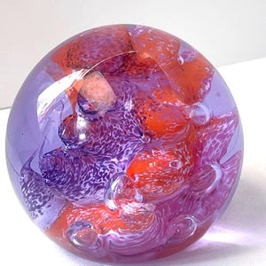 May include: A spherical glass paperweight featuring a clear outer layer encasing a vibrant interior of swirling purple and orange patterns. The design includes intricate white speckled details and small trapped air bubbles throughout the glass sphere.
