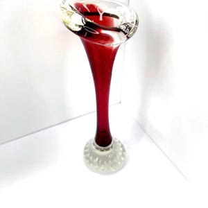 Peut inclure: Un vase en verre haut et élancé avec un corps en forme de trompette rouge profond qui s'évase vers le haut. Le bord est en verre transparent et la base est un piédestal rond en verre transparent avec un motif texturé en forme de bulles.