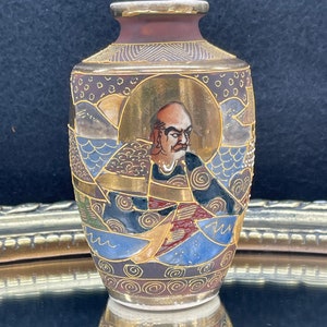 Puede incluir: Un peque&ntilde;o jarr&oacute;n de cer&aacute;mica pintado a mano con un dise&ntilde;o dorado, marr&oacute;n, azul y blanco. El jarr&oacute;n presenta una representaci&oacute;n de un hombre con una expresi&oacute;n severa.