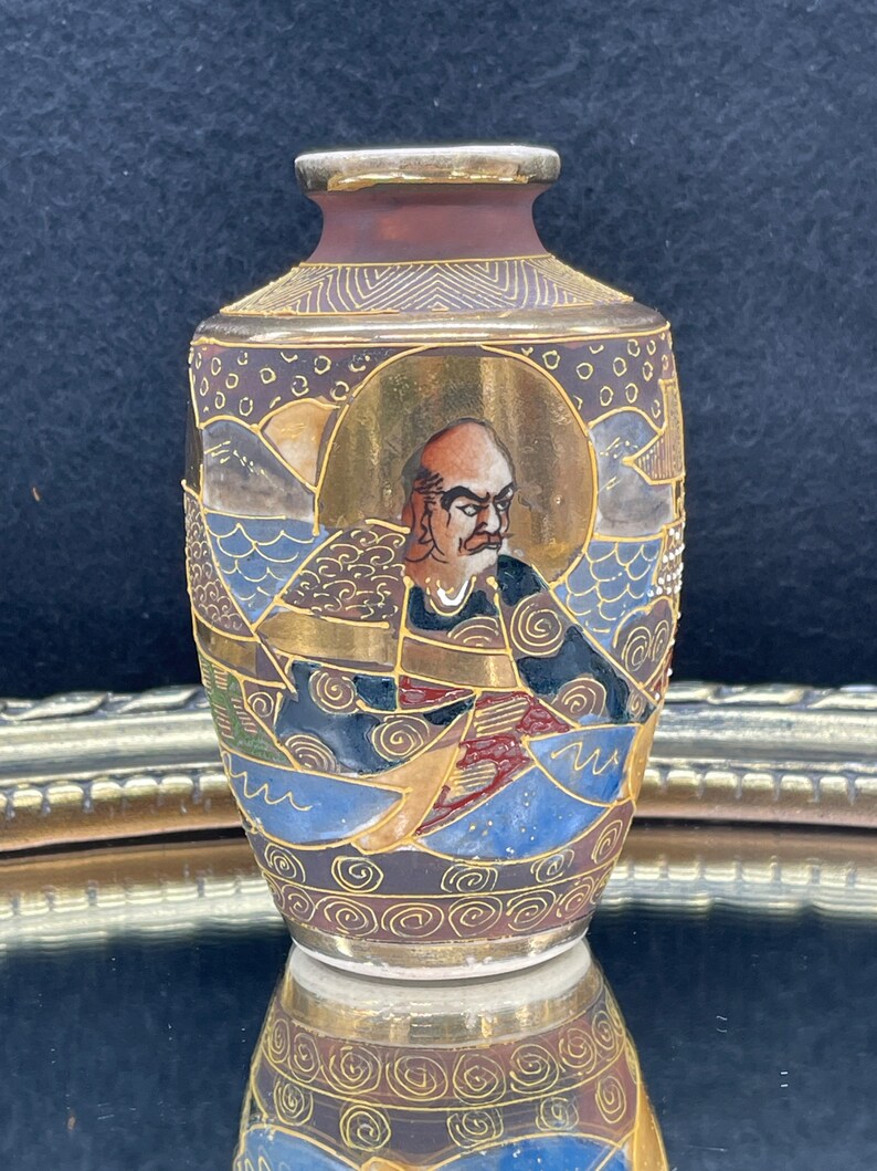 Puede incluir: Un peque&ntilde;o jarr&oacute;n de cer&aacute;mica pintado a mano con un dise&ntilde;o dorado, marr&oacute;n, azul y blanco. El jarr&oacute;n presenta una representaci&oacute;n de un hombre con una expresi&oacute;n severa.