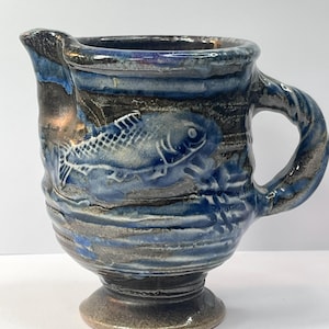 May include: Handmade ceramic pitcher with a blue and gray glaze. The pitcher features a raised relief design of a fish on the side. It has a handle and a spout for pouring. The base is a pedestal shape.