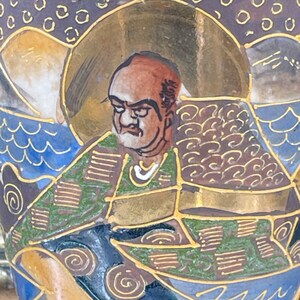 Puede incluir: Un jarr&oacute;n de cer&aacute;mica con un dise&ntilde;o dorado, azul y verde. El jarr&oacute;n presenta una imagen pintada de un hombre con barba y una t&uacute;nica estampada. El hombre est&aacute; rodeado de olas y nubes estilizadas.
