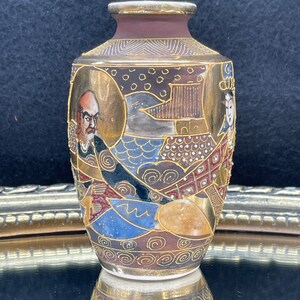 Puede incluir: Un peque&ntilde;o jarr&oacute;n de cer&aacute;mica decorativo con base marr&oacute;n y dise&ntilde;os intrincados en oro y varios colores. El jarr&oacute;n presenta una escena de inspiraci&oacute;n japonesa con figuras y patrones geom&eacute;tricos.