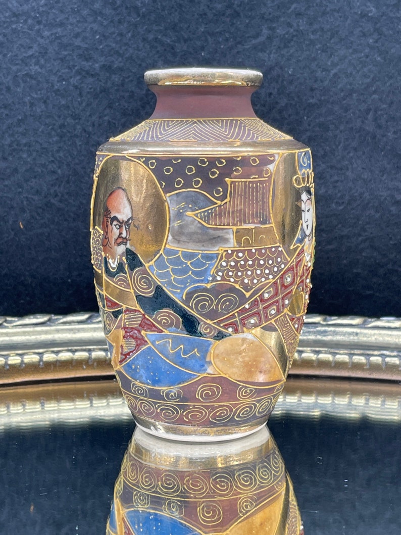 Puede incluir: Un peque&ntilde;o jarr&oacute;n de cer&aacute;mica decorativo con base marr&oacute;n y dise&ntilde;os intrincados en oro y varios colores. El jarr&oacute;n presenta una escena de inspiraci&oacute;n japonesa con figuras y patrones geom&eacute;tricos.