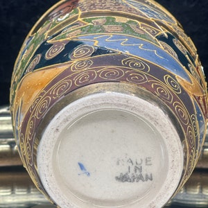 Puede incluir: Un peque&ntilde;o jarr&oacute;n redondo de cer&aacute;mica con un dise&ntilde;o floral dorado y marr&oacute;n. El jarr&oacute;n tiene una base blanca con las palabras "Made in Japan" impresas en &eacute;l.