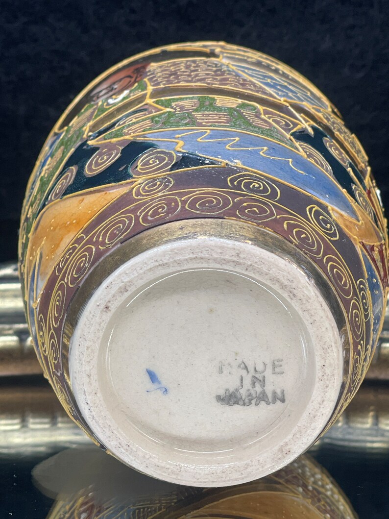 Puede incluir: Un peque&ntilde;o jarr&oacute;n redondo de cer&aacute;mica con un dise&ntilde;o floral dorado y marr&oacute;n. El jarr&oacute;n tiene una base blanca con las palabras "Made in Japan" impresas en &eacute;l.