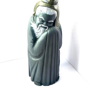May include: A dark gray ceramic figurine of an elderly person with a long white beard and hair, wearing a hooded robe. The figure has closed eyes and a serene expression, with a smooth matte finish and subtle highlights on the hair and beard.