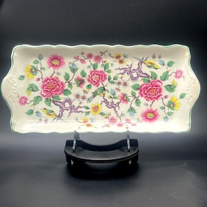 Könnte beinhalten: Ein rechteckiges Serviertablett aus Keramik mit gewelltem Rand und cremefarbener Basis. Die Oberfläche zeigt ein buntes Blumenmuster mit leuchtend rosa Pfingstrosen, gelben Blüten und kleinen Vögeln auf violetten Zweigen mit grünen Blättern.