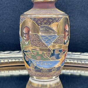 Puede incluir: Un peque&ntilde;o jarr&oacute;n de cer&aacute;mica marr&oacute;n y dorado con un dise&ntilde;o detallado de figuras, olas y monta&ntilde;as. El jarr&oacute;n est&aacute; decorado con pintura dorada y tiene un acabado brillante.