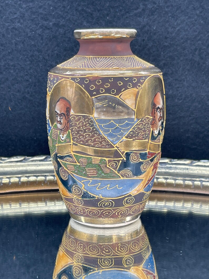 Puede incluir: Un peque&ntilde;o jarr&oacute;n de cer&aacute;mica marr&oacute;n y dorado con un dise&ntilde;o detallado de figuras, olas y monta&ntilde;as. El jarr&oacute;n est&aacute; decorado con pintura dorada y tiene un acabado brillante.