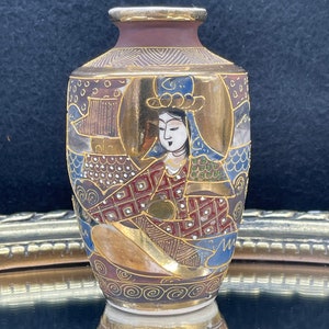 Puede incluir: Un peque&ntilde;o jarr&oacute;n de cer&aacute;mica pintado a mano con un dise&ntilde;o dorado y rojo. El jarr&oacute;n presenta una imagen detallada de una mujer con ropa tradicional japonesa.