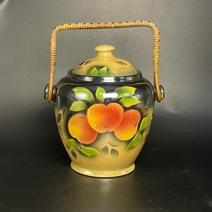 May include: A ceramic jar with a lid and a woven rattan handle. The jar features a hand-painted design of three orange peaches with green leaves against a black and tan background. The lid is tan with a small knob handle at the centre.