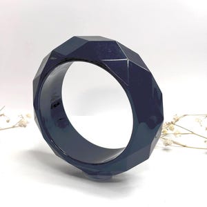 Könnte beinhalten: Ein dunkelblaues, facettiertes Armreif-Armband. Das Armband hat ein geometrisches Design mit mehreren flachen Oberflächen. Der Armreif besteht aus glänzendem Material und ist vor einem weißen Hintergrund platziert.