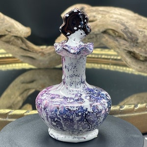 Pode incluir: Um pequeno frasco de cerâmica branca com um design marmorizado roxo e preto. O frasco tem uma tampa decorativa preta e branca.