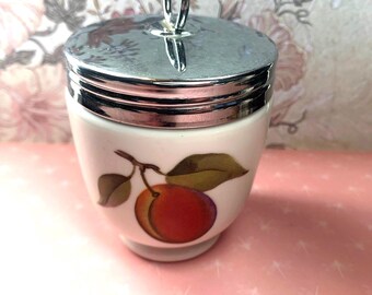May include: A white ceramic egg cup with a silver lid and a single peach design. The lid has a silver ring handle.
