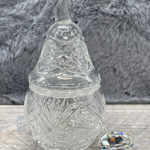May include: A clear glass pear-shaped jar with a lid and a crystal-like embellishment. The jar has a cut-glass design.