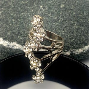 May include: A silver ring with a design of four white flower accents, each with a pearl center and clear rhinestones.