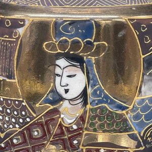 Puede incluir: Un jarr&oacute;n de cer&aacute;mica japon&eacute;s vintage con fondo dorado y marr&oacute;n. El jarr&oacute;n est&aacute; decorado con una escena colorida de una mujer con una t&uacute;nica azul y dorada. La cara de la mujer est&aacute; pintada en detalle con ojos negros y una boca roja.