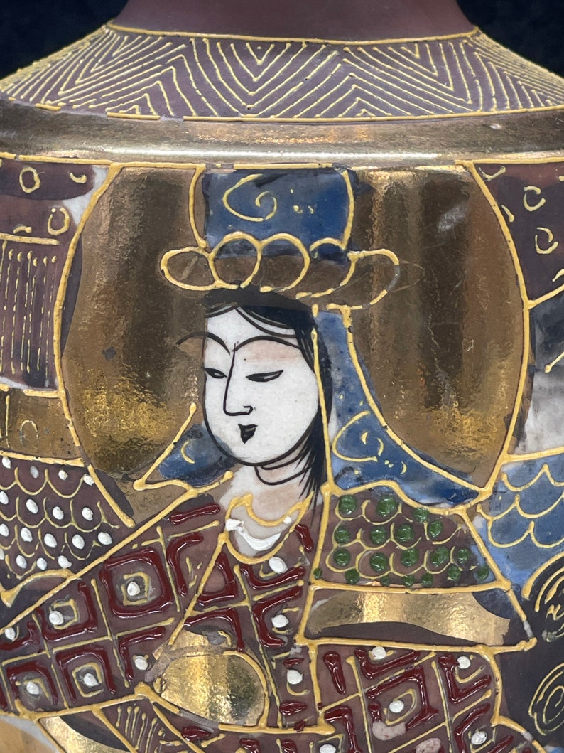 Puede incluir: Un jarr&oacute;n de cer&aacute;mica japon&eacute;s vintage con fondo dorado y marr&oacute;n. El jarr&oacute;n est&aacute; decorado con una escena colorida de una mujer con una t&uacute;nica azul y dorada. La cara de la mujer est&aacute; pintada en detalle con ojos negros y una boca roja.