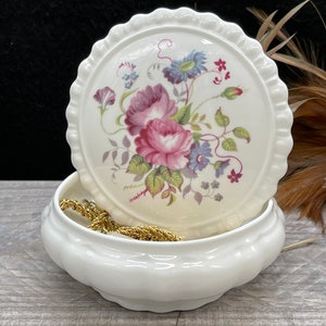 May include: White porcelain trinket box with a floral design on the lid. The lid has a scalloped edge. The box is open and a gold chain is inside.