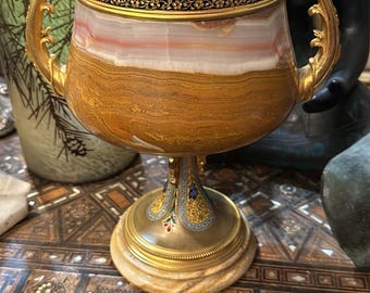 May include: A gold and onyx vase with intricate enamel detailing. The vase has two handles and a round base. The enamel detailing features floral and geometric patterns in shades of blue, red, and gold.
