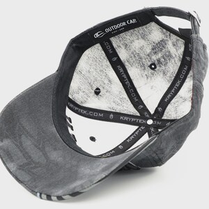 May include: A gray baseball cap with a curved brim, viewed from the underside. The interior is white with a camouflage pattern. The cap has the text "OUTDOOR CAP" and "KRYPTEK.COM" printed on the inside.