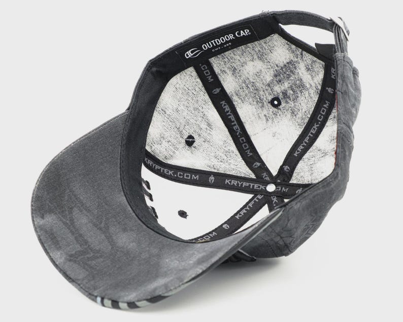May include: A gray baseball cap with a curved brim, viewed from the underside. The interior is white with a camouflage pattern. The cap has the text "OUTDOOR CAP" and "KRYPTEK.COM" printed on the inside.