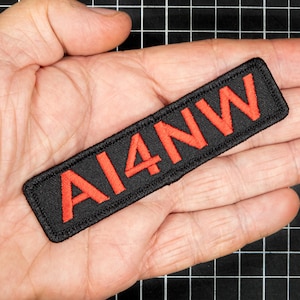 May include: A black rectangular patch with the embroidered text "AI4NW" in red. The patch is held in a person's hand, and the background is a black grid.