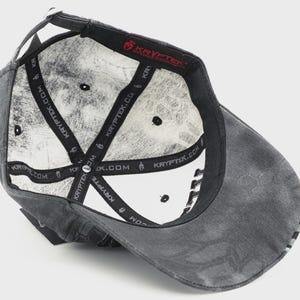 May include: A gray baseball cap with a distressed finish, viewed from the inside. The interior features a white pattern and black straps with "KRYPTEK.COM" printed on them. The cap has a curved brim and a small, black button on top.